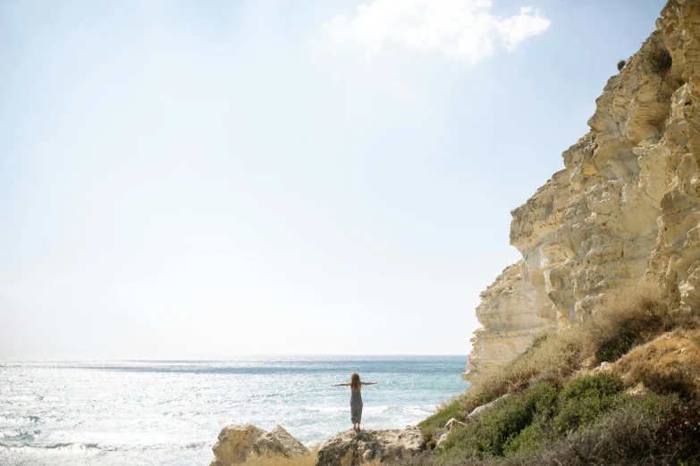 a man standing on top of a cliff next to the ocean, by Will Ellis, unsplash, minimalism, margot robbie on the beach, cyprus, clear blue skies, photograph of april