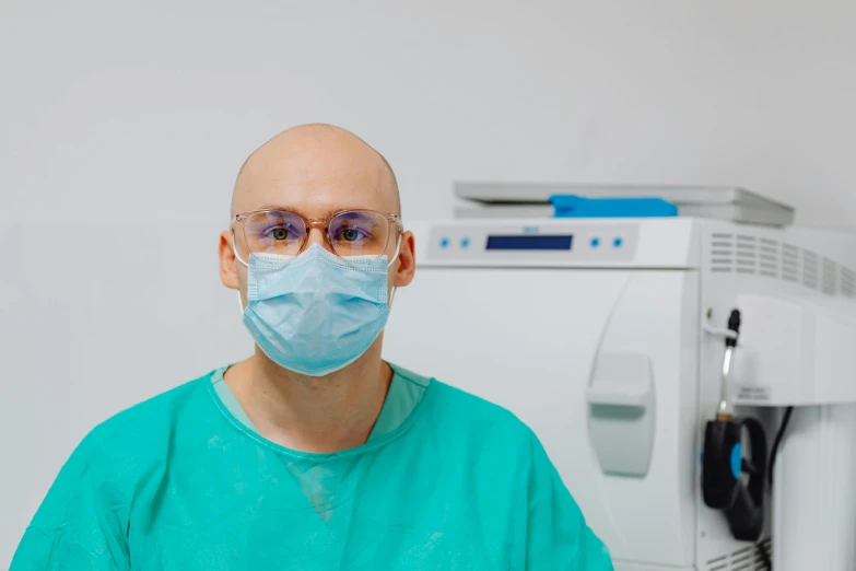 a man wearing a surgical mask standing in front of a machine, a picture, by Adam Marczyński, portrait of bald, dentist, 8k octan photo, professional profile picture