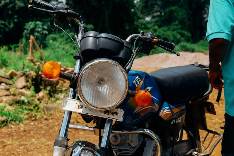 a man standing next to a motorcycle on a dirt road, pexels contest winner, photorealism, round headlights, sri lanka, close up details, blue and orange rim lights