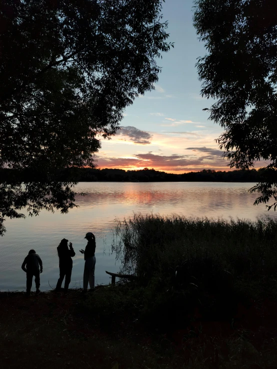 a group of people standing next to a body of water, sundown, near a lake, evening time, from the distance