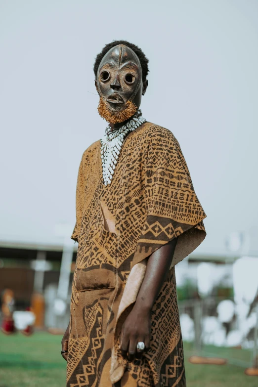 a man wearing a mask standing in a field, trending on unsplash, afrofuturism, wearing an african dress, brown body, evil looking, market