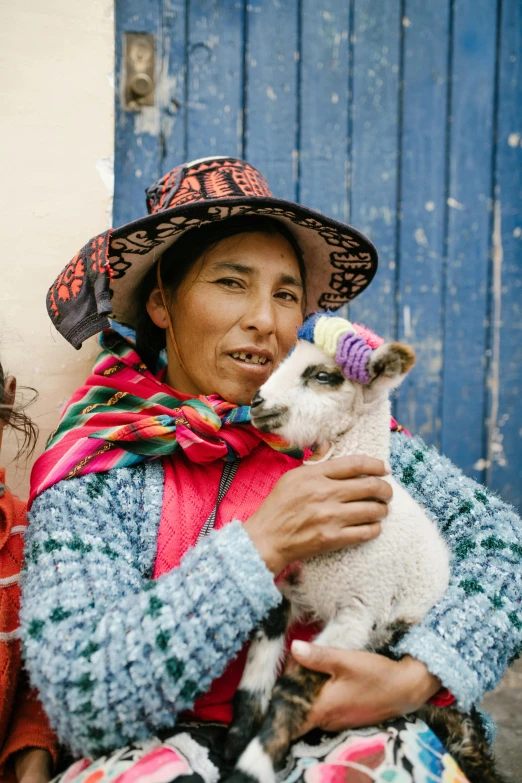 a woman holding a baby sheep in front of a blue door, inspired by Steve McCurry, trending on unsplash, bolivian cholitas, animal hat, lesbians, square