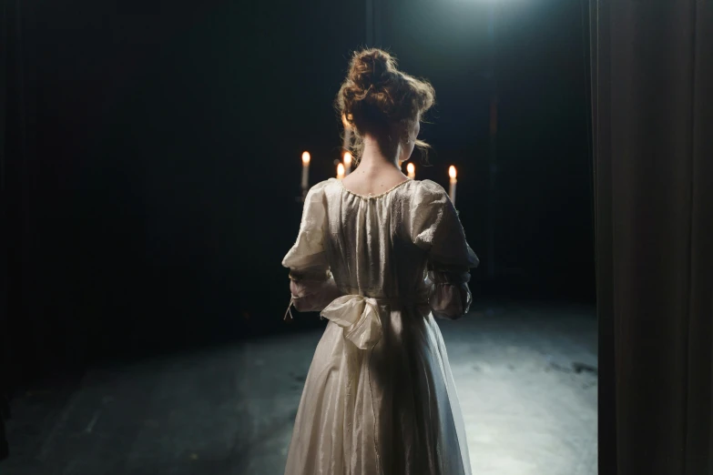 a woman in a white dress standing in a dark room, lit with candles, actors, back facing, ( ( theatrical ) )