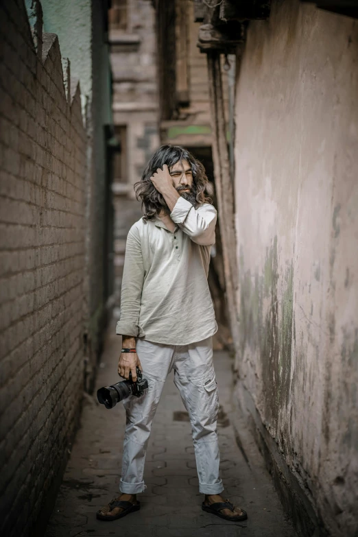 a man standing in an alley talking on a cell phone, inspired by Steve McCurry, pexels contest winner, long messy hair, holding a dslr camera, dressed in a white t shirt, nepal