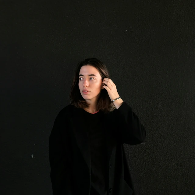 a woman standing in front of a black wall talking on a cell phone, an album cover, by Alejandro Obregón, pexels contest winner, wearing a black sweater, androgynous person, shoulder-length hair, human staring blankly ahead