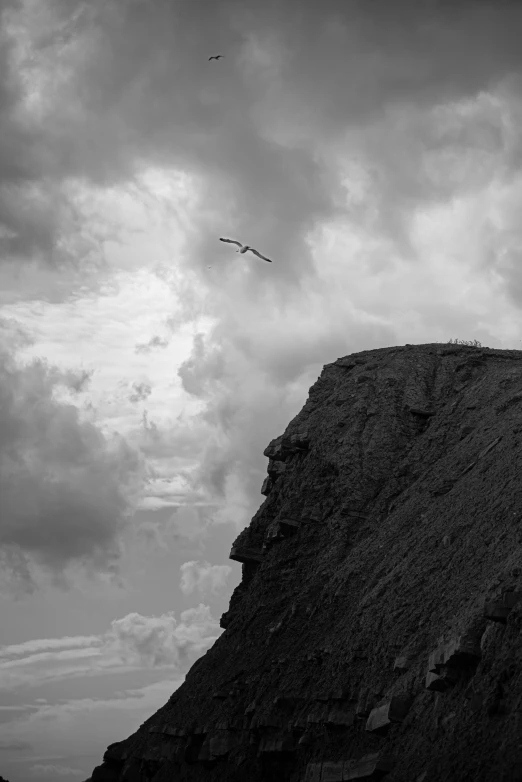 a black and white photo of a bird flying over a cliff, by Altichiero, dramatic skies, faroe, surrealism - n 9, heads