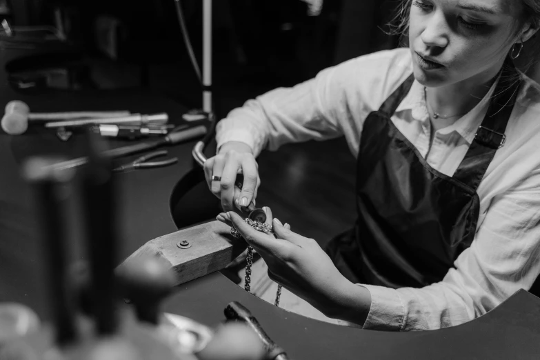 a woman in an apron working on a piece of wood, a black and white photo, by Emma Andijewska, pexels contest winner, wearing elegant jewellery, lawther sit at table playing dnd, customers, leather jewelry