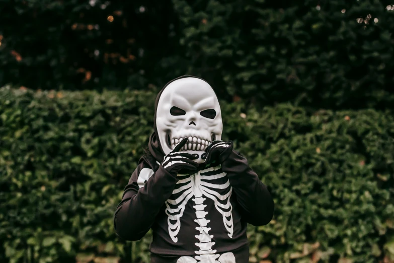 a person wearing a skeleton mask standing in front of a hedge, a cartoon, pexels contest winner, little kid, white bones, balaclava, dark and white