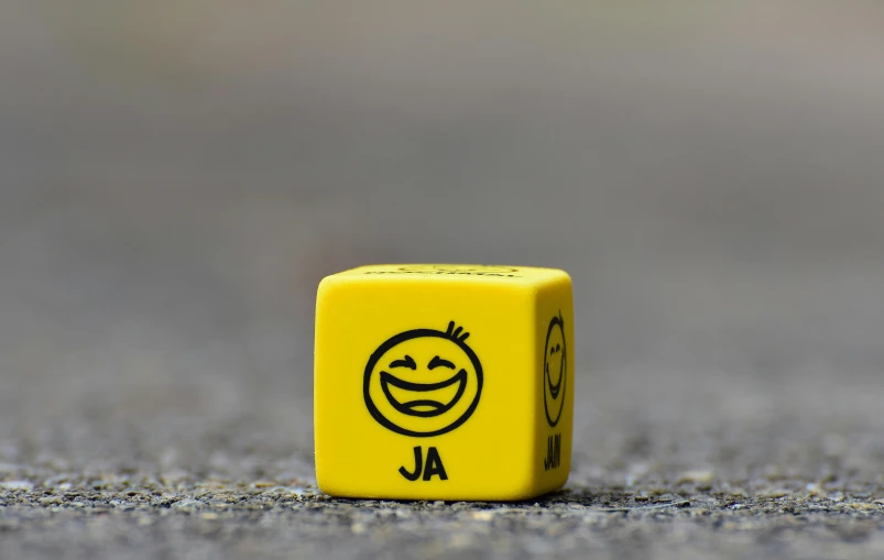 a yellow dice with a smiley face drawn on it, a picture, by Joze Ciuha, unsplash, square masculine jaw, on a road, ja mong, smiling like a jerk