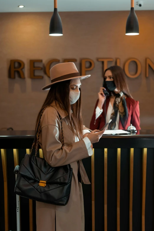 a woman wearing a face mask standing in front of a reception counter, pexels, renaissance, caracter with brown hat, checking her phone, hotel, two women
