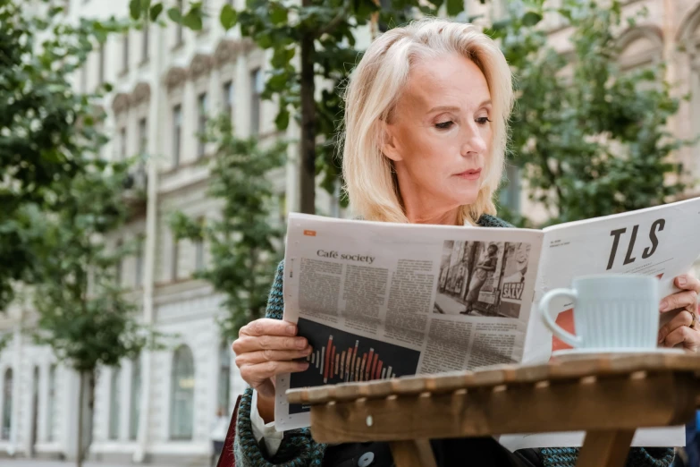 a woman sitting at a table reading a newspaper, a photo, by Emma Andijewska, pexels contest winner, blonde swedish woman, in the city, middle - age, gif