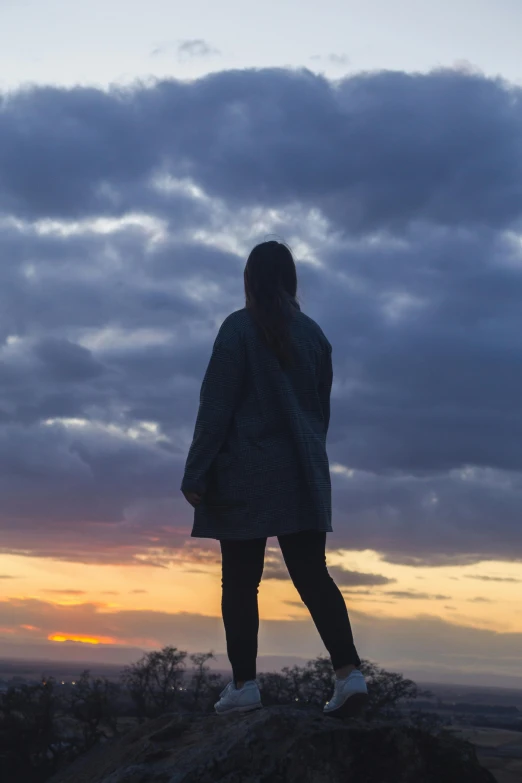 a person standing on top of a mountain at sunset, backlit girl in raincoat, gazing off into the horizon, grey, teenage girl