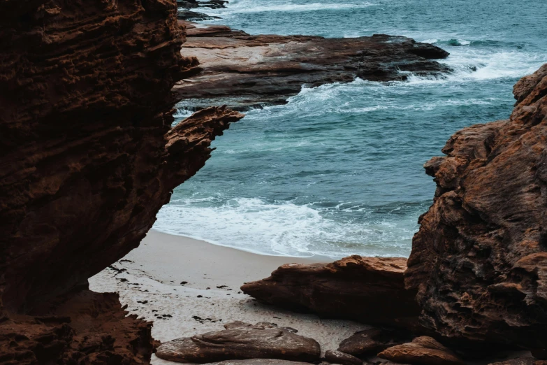 a man standing on top of a sandy beach next to the ocean, pexels contest winner, australian tonalism, natural cave wall, red sand, turquoise water, rocks coming out of the ground