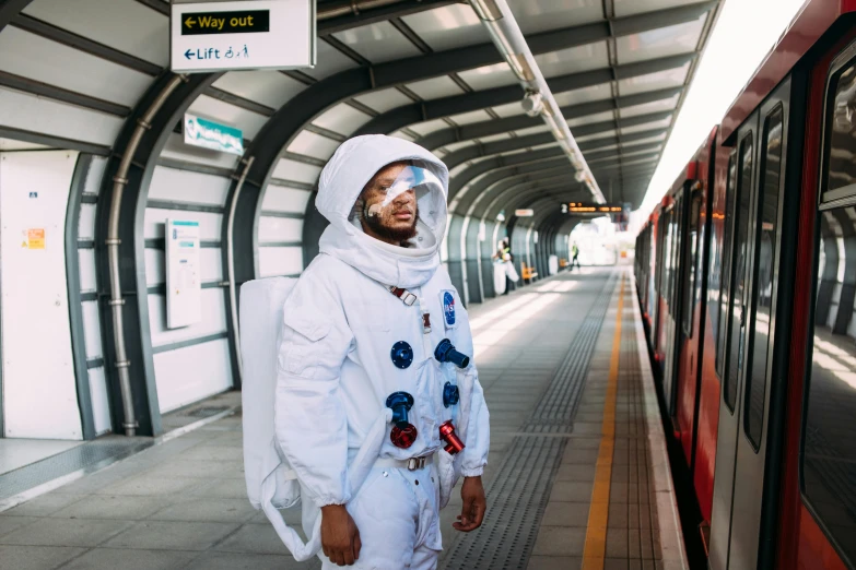 a man in a space suit standing next to a train, by Cosmo Alexander, pexels contest winner, afrofuturism, in london, childish gambino, lunar themed attire, dressed a long white