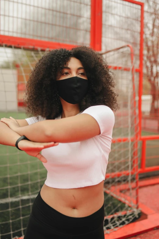 a woman standing in front of a soccer goal wearing a face mask, trending on pexels, black bandage on arms, wearing a crop top, infp young woman, square