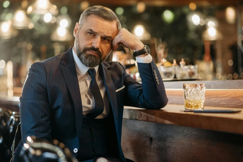 a man in a suit sitting at a bar, inspired by Edi Rama, well trimmed beard, whisky, 4 5 yo, thumbnail