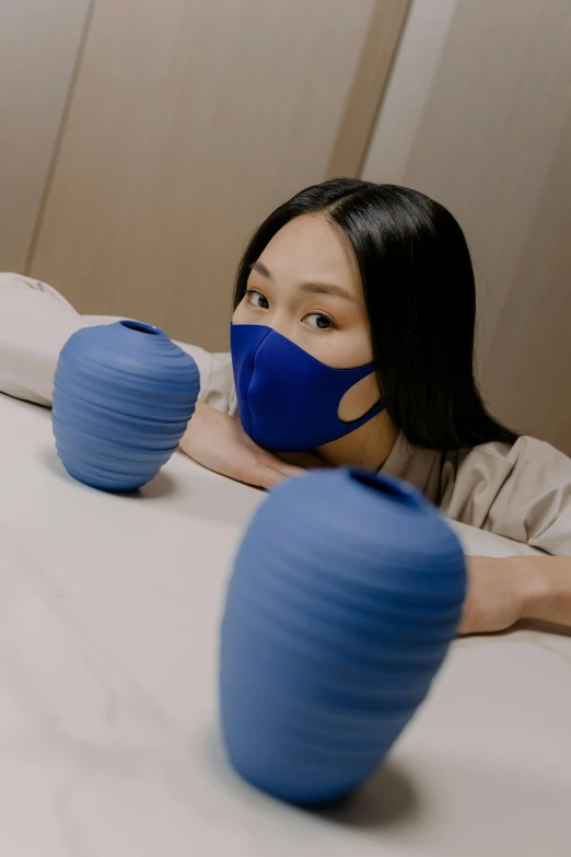 a woman sitting at a table with blue vases, inspired by Yu Zhiding, happening, wearing facemask, bed, silicone cover, punching