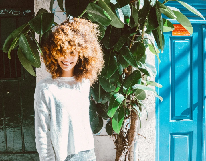 a woman standing in front of a blue door, by Julia Pishtar, pexels contest winner, brown curly hair, next to a plant, wearing a white sweater, happy girl