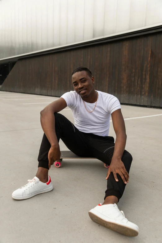 a man sitting on a skateboard in front of a building, jaylen brown, profile image, in front of white back drop, wearing pants and a t-shirt