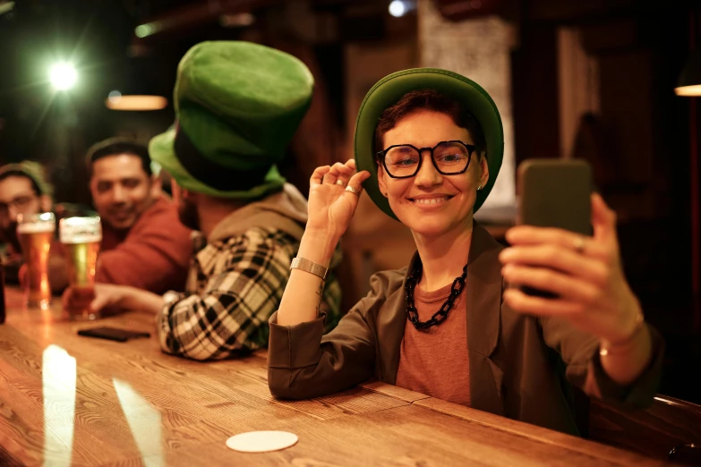 a woman sitting at a bar taking a picture of herself, pexels contest winner, green hat, irish, square rimmed glasses, college party