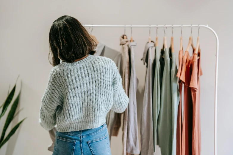 a woman standing in front of a rack of clothes, by Nicolette Macnamara, trending on pexels, standing with her back to us, wearing a sweater, pastel colour palette, sustainable materials