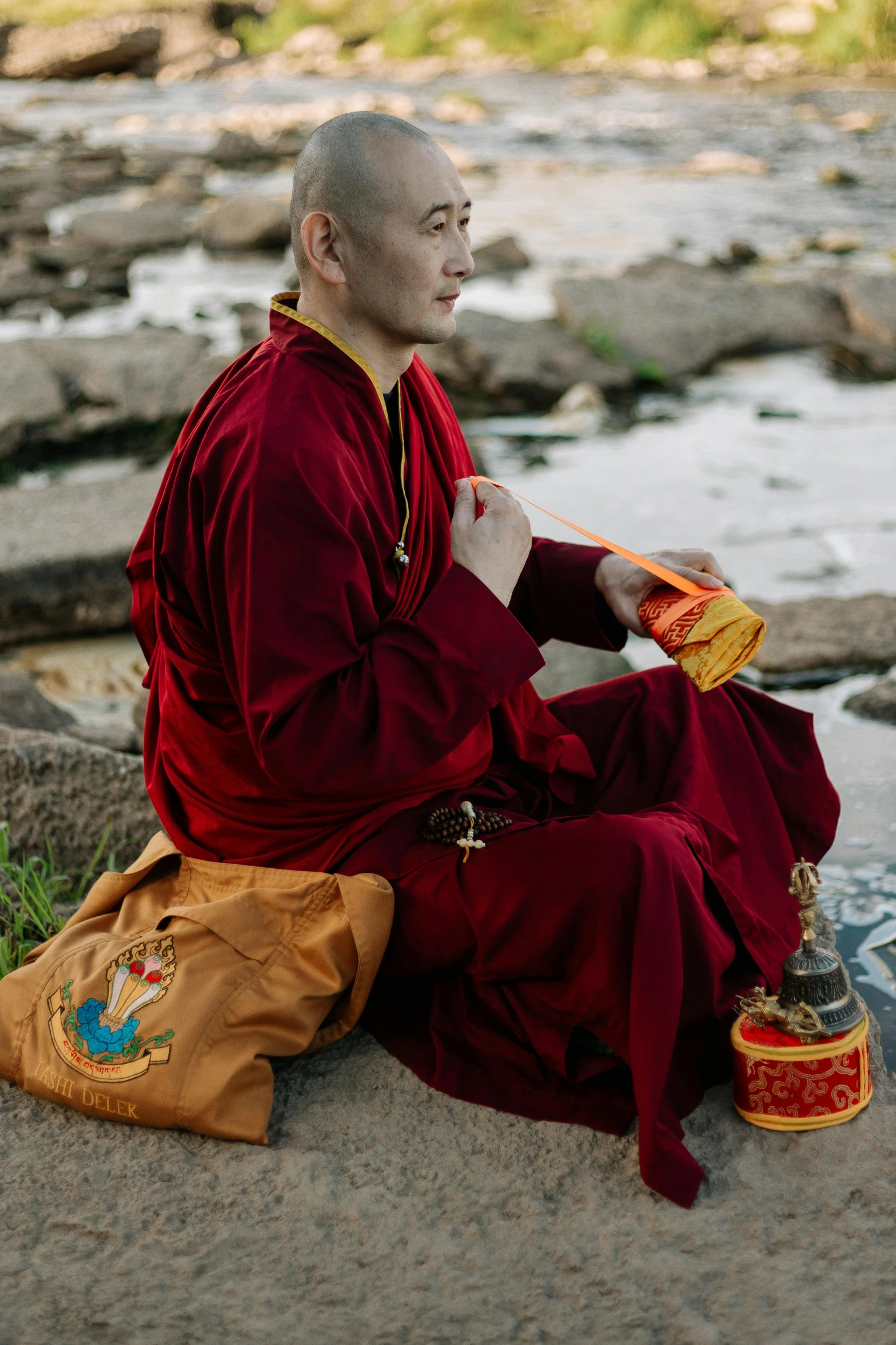 a man sitting on top of a rock next to a river, a portrait, inspired by Ma Quan, unsplash, wearing gilded red robes, holding a gold bag, ukrainian monk, profile image