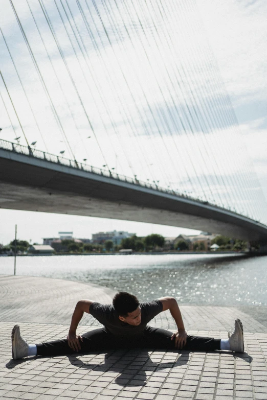 a man laying on the ground in front of a bridge, by Yosa Buson, unsplash, bangkok, doing splits and stretching, at the waterside, on the bow