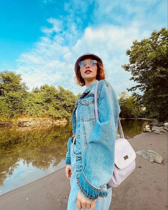 a woman standing next to a body of water, featured on instagram, wearing a jeans jackets, beret and sunglasses, bag over the waist, 🤬 🤮 💕 🎀