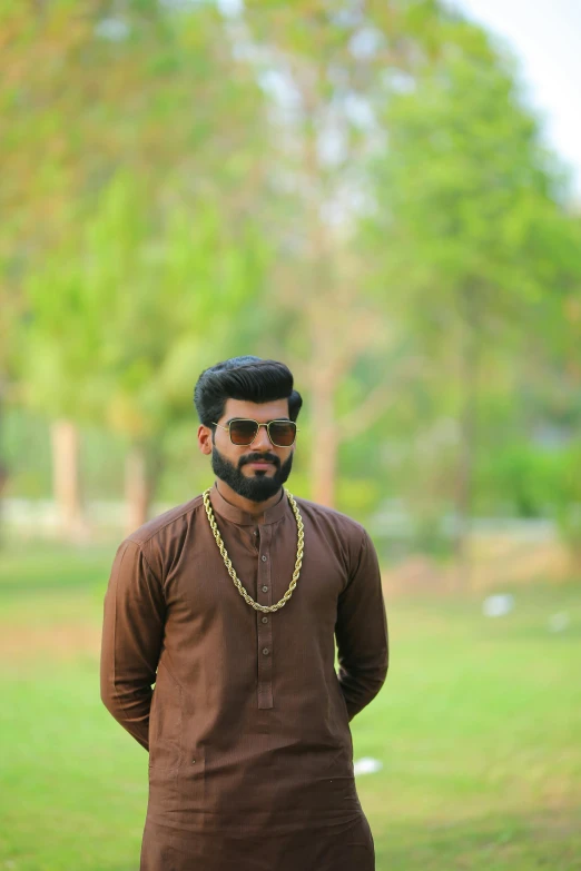 a man standing on top of a lush green field, by Max Dauthendey, pexels contest winner, hurufiyya, wearing a gold chain, brown beard, at college, wearing gold jewellery
