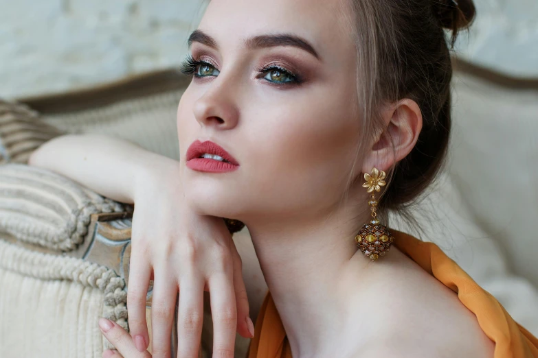 a woman in an orange dress sitting on a couch, a portrait, trending on pixabay, wearing ornate earrings, soft pale golden skin, with professional makeup, topaz and other jewellaries