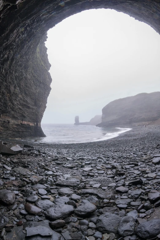 a cave filled with lots of rocks next to the ocean, a picture, inspired by Johan Christian Dahl, pexels contest winner, romanticism, under a gray foggy sky, icelandic valley, deserted sand, bald head