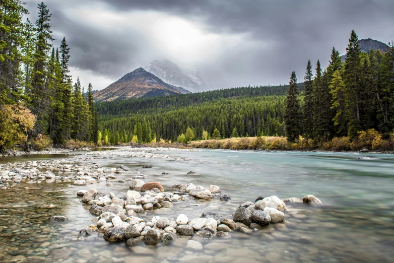 a river running through a forest filled with rocks, inspired by James Pittendrigh MacGillivray, unsplash contest winner, rocky mountains in background, grey cloudy skies, conde nast traveler photo, banff national park