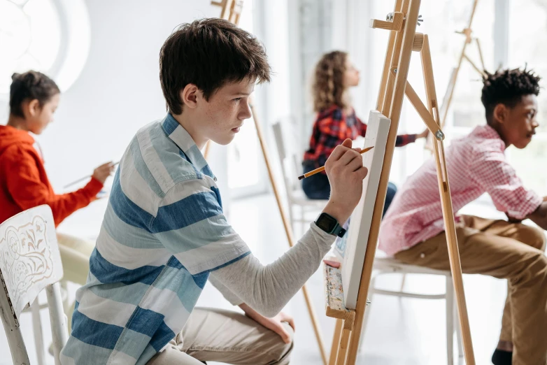 a man sitting on a chair in front of a painting easel, inspired by Arthur Streeton, pexels contest winner, academic art, teenage boy, children drawing with pencils, looking across the shoulder, on a canva