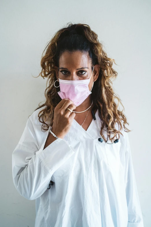 a woman wearing a face mask in front of a white wall, happening, pastel pink robes, lab coat and tee shirt, hand over mouth, r&b