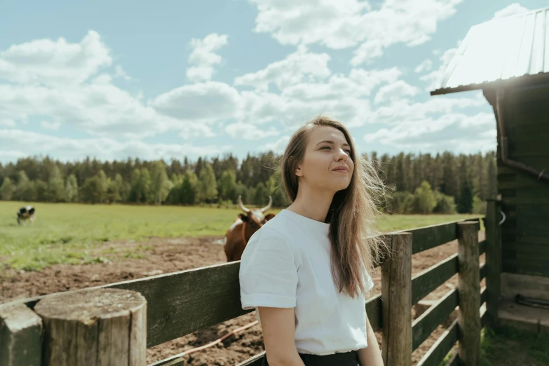 a woman standing in front of a fence next to a cow, by Emma Andijewska, pexels contest winner, greta thunberg smiling, looking at the sky, dressed in a white t shirt, jenni pasanen