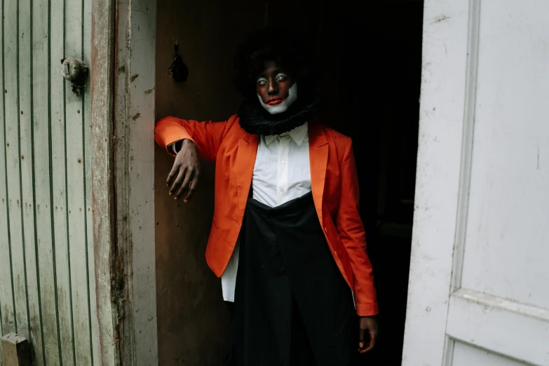 a man dressed as a clown standing in a doorway, an album cover, pexels contest winner, man is with black skin, sarah cliff, black and orange coat, lachlan bailey