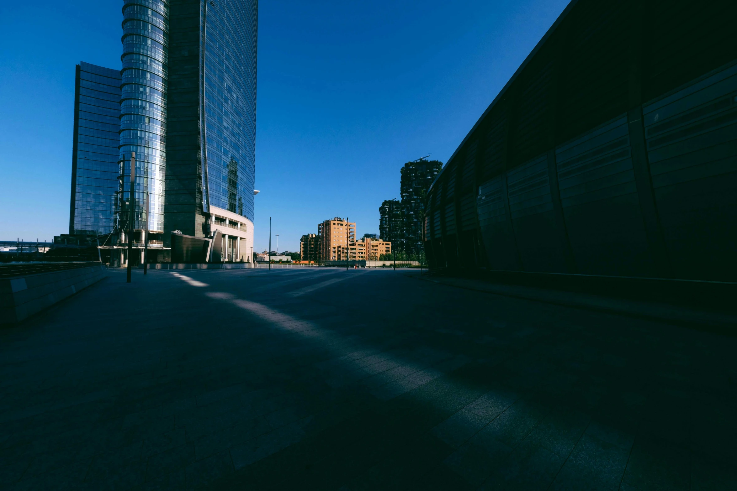 a man riding a skateboard down a street next to tall buildings, a picture, unsplash contest winner, minimalism, cloudless sky, madrid. extreme long shot, harsh shadows and reflections, location [ chicago ( alley ) ]