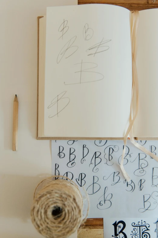 an open book sitting on top of a table next to a spool of twine of twine, by Lucette Barker, letterism, monogram, in a white boho style studio, children drawing with pencils, thumbnail