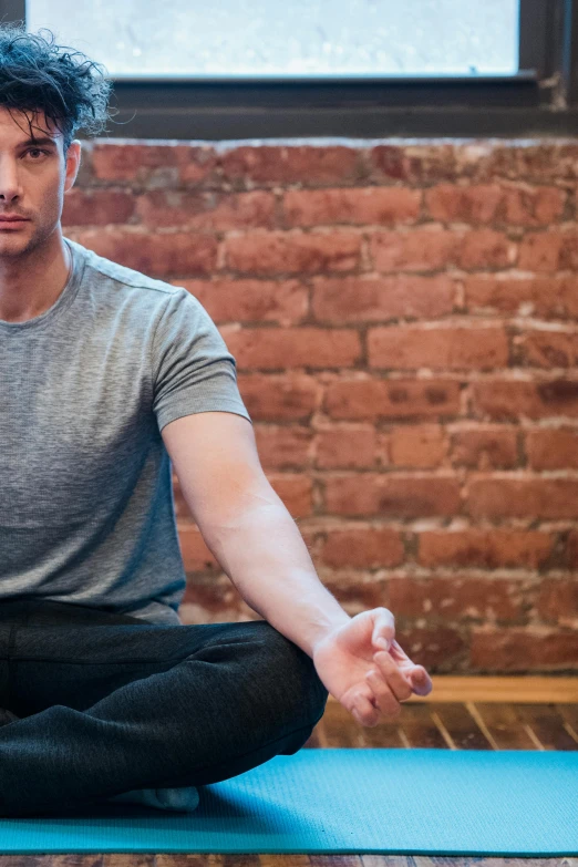 a man sitting on a yoga mat in front of a brick wall, a portrait, unsplash, insectile forearms folded, panoramic shot, square, kyle mclaughlin