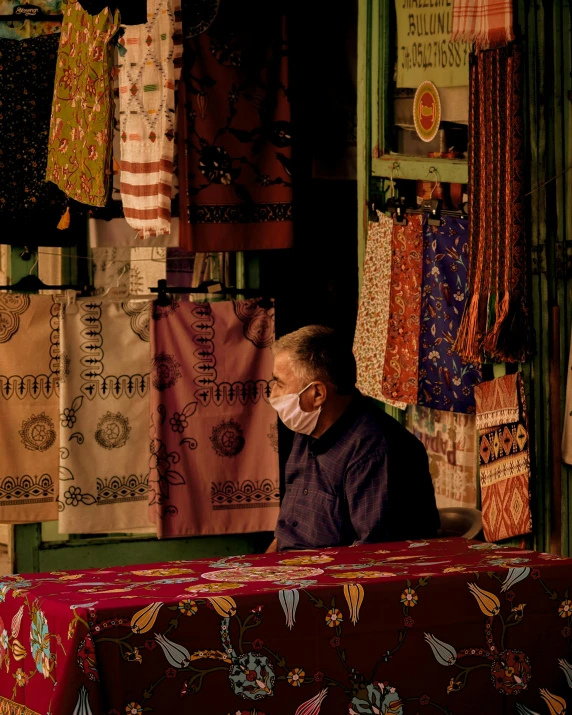 a man sitting at a table with a mask on, a silk screen, trending on unsplash, cloisonnism, old shops, several layers of fabric, jakarta, night photo