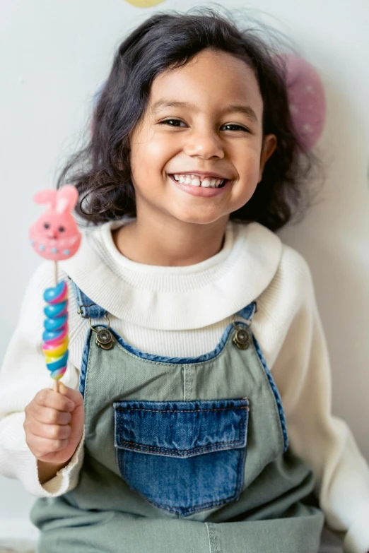 a little girl sitting on the floor holding a lollipop lollipop lollipop lollipop lollipop lollipop lo, pexels contest winner, wearing blue jean overalls, she is smiling and happy, easter, gummy worms