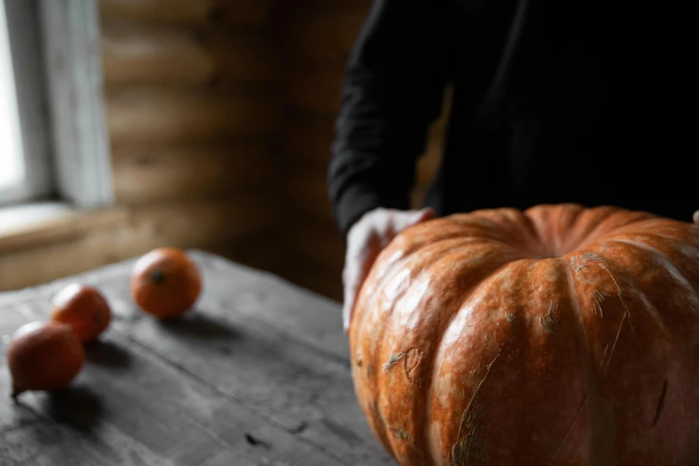 a person holding a pumpkin on top of a wooden table, by Emma Andijewska, unsplash, rustic yet enormous scp (secure, grey orange, carving, very large bosum