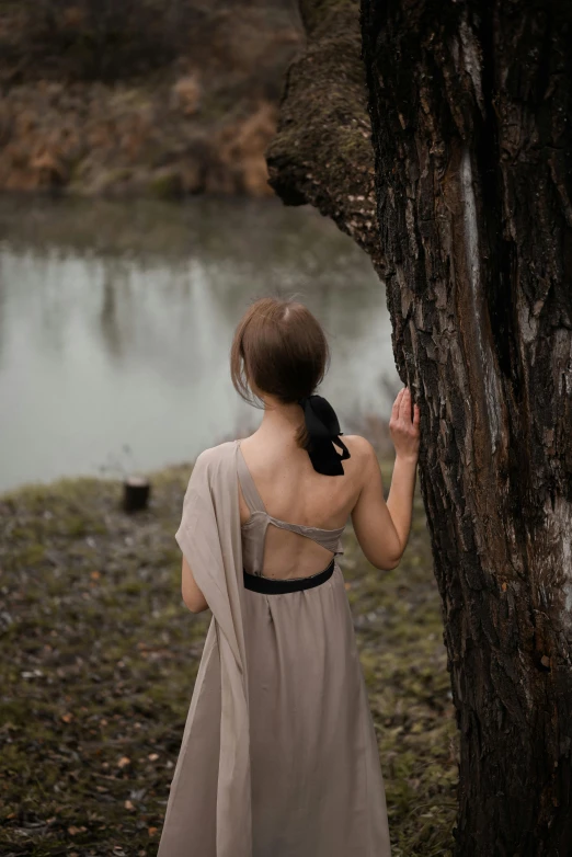 a woman standing next to a tree near a body of water, inspired by Camille Corot, unsplash, renaissance, bare back, soft silk dress, low quality photo, grey