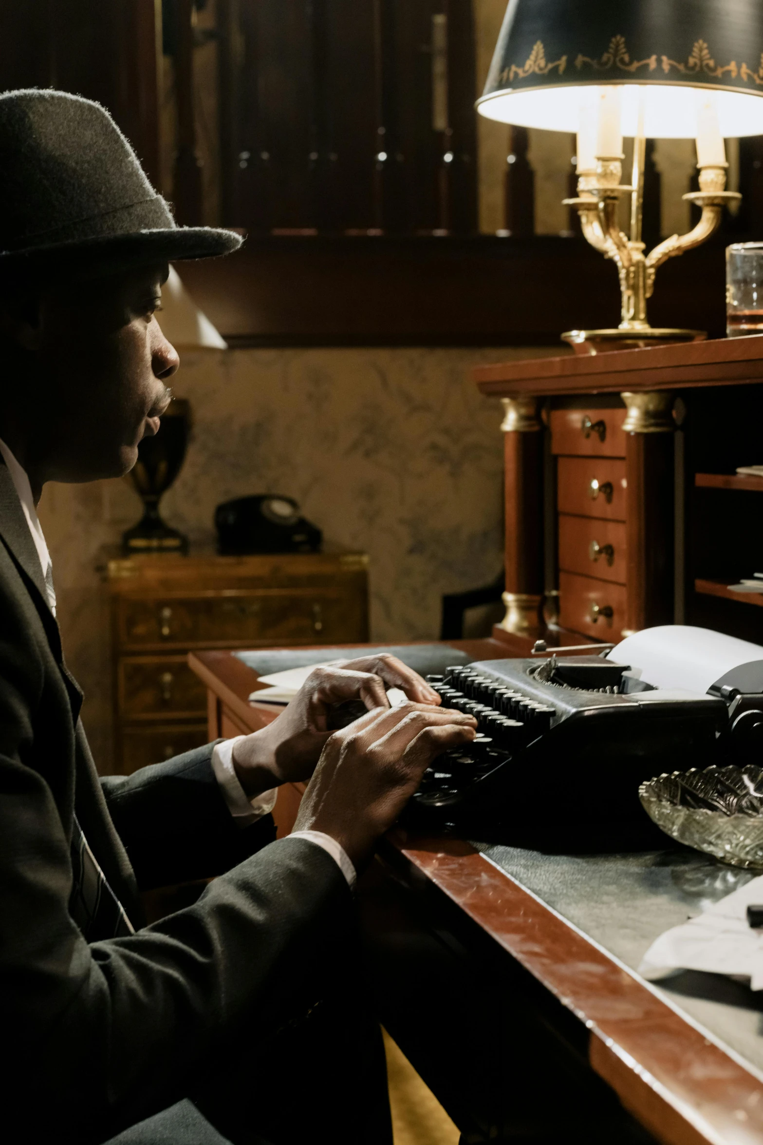 a man sitting at a desk typing on a typewriter, inspired by Maxwell Bates, harlem renaissance, cinematic lighting + masterpiece, a suited man in a hat, historical setting, full - shot