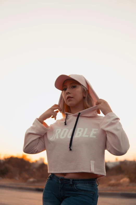 a woman standing in the middle of a road wearing a pink hoodie, an album cover, by Robbie Trevino, trending on pexels, wearing a baseball cap, punchable expression, sundown, dribble popular
