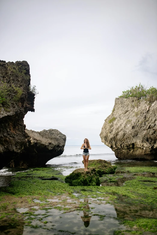 a woman standing on a rock in front of a body of water, bali, rock arches, grass and rocks, beaching