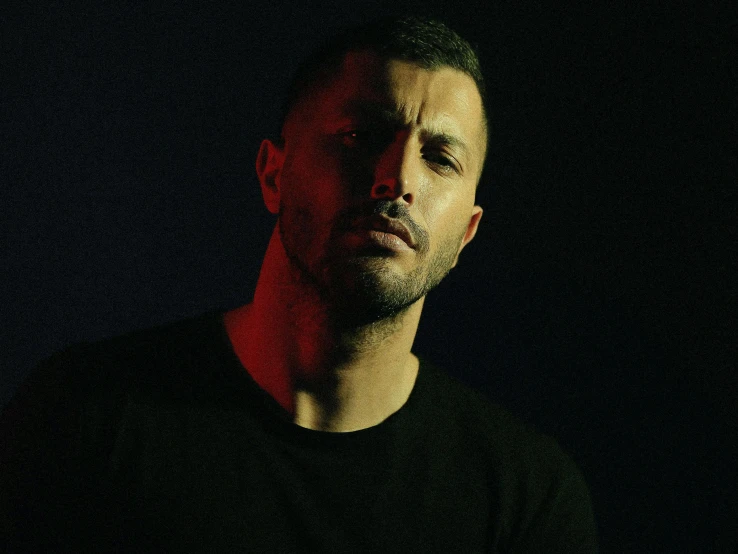 a man standing in front of a black background, an album cover, inspired by Nadim Karam, pexels, les nabis, looking serious, gal yosef, light falling on face, photo from a promo shoot
