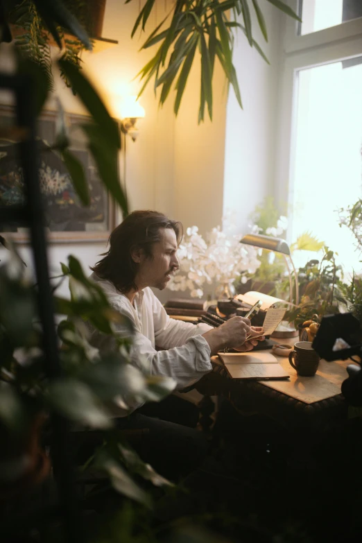 a man sitting at a desk in front of a window, by Else Alfelt, unsplash, romanticism, cats and plants, soft light.4k, frank dillane, sitting on a mocha-colored table