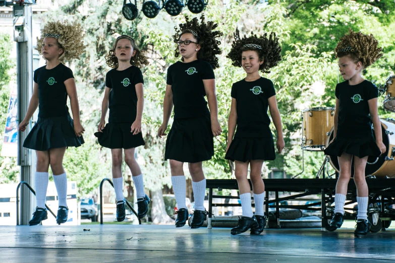 a group of young girls standing on top of a stage, green and black color scheme, trees swaying to the beat, wearing a kilt, 🌻🎹🎼