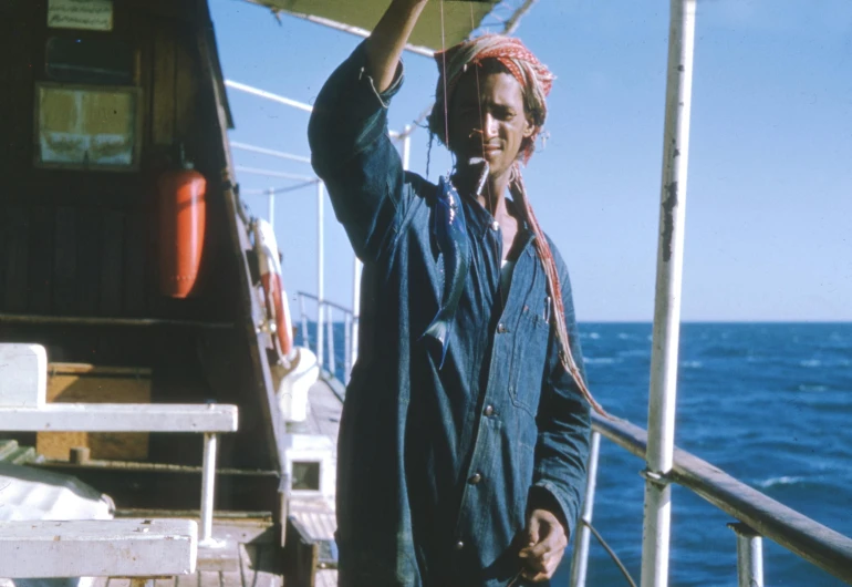 a man standing on top of a boat next to the ocean, a colorized photo, holding an fish in his hand, still from a terence malik film, australian, arthur clarke